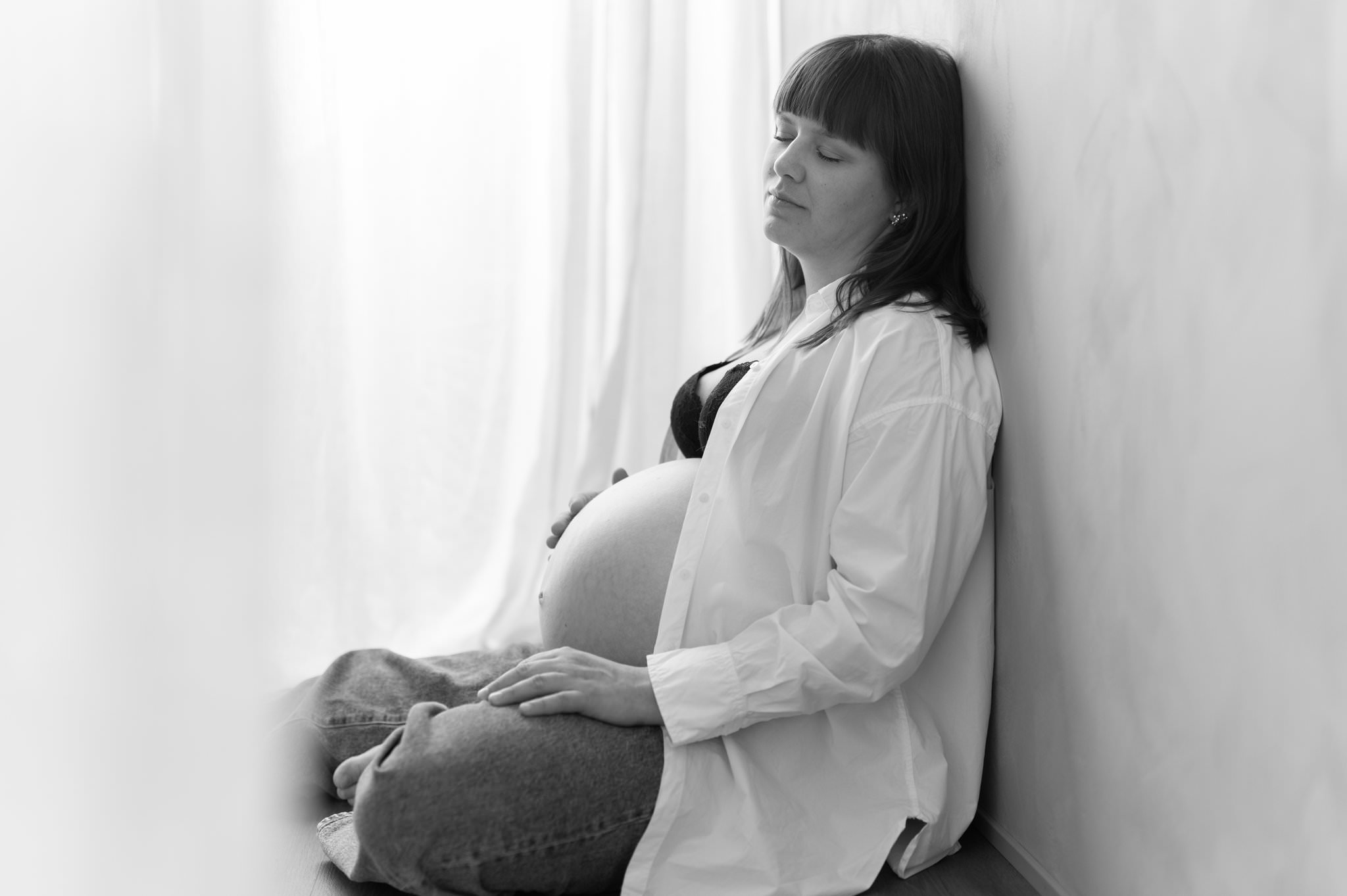 Avslappnad gravidfoto i mysig studio i Skåne. Kvinna sitter med ryggen mot en vägg och blundar. 