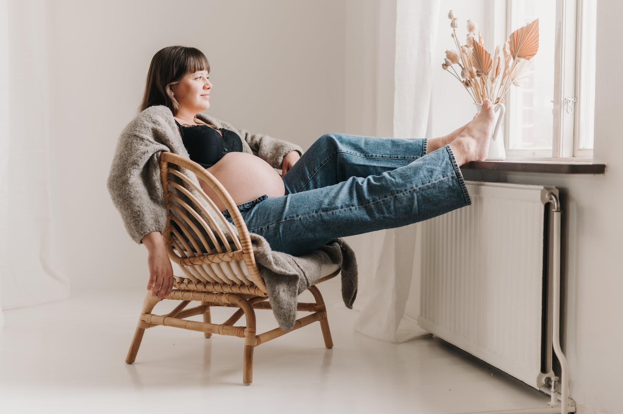 Gravidfotografering i studio där kvinnan sitter i en stol med fötterna på fönsterkarmen och tittar ut genom fönstret. 